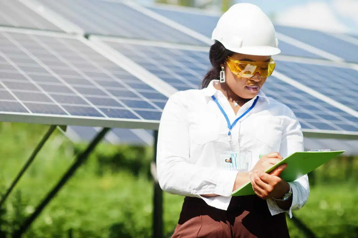 Mujer ingeniera trabajando en un parque solar, tomando notas con paneles fotovoltaicos de fondo.