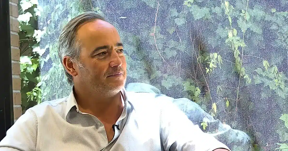 Directivo del sector energético en conversación, camisa celeste, gesto sereno; fondo vegetal en interior.