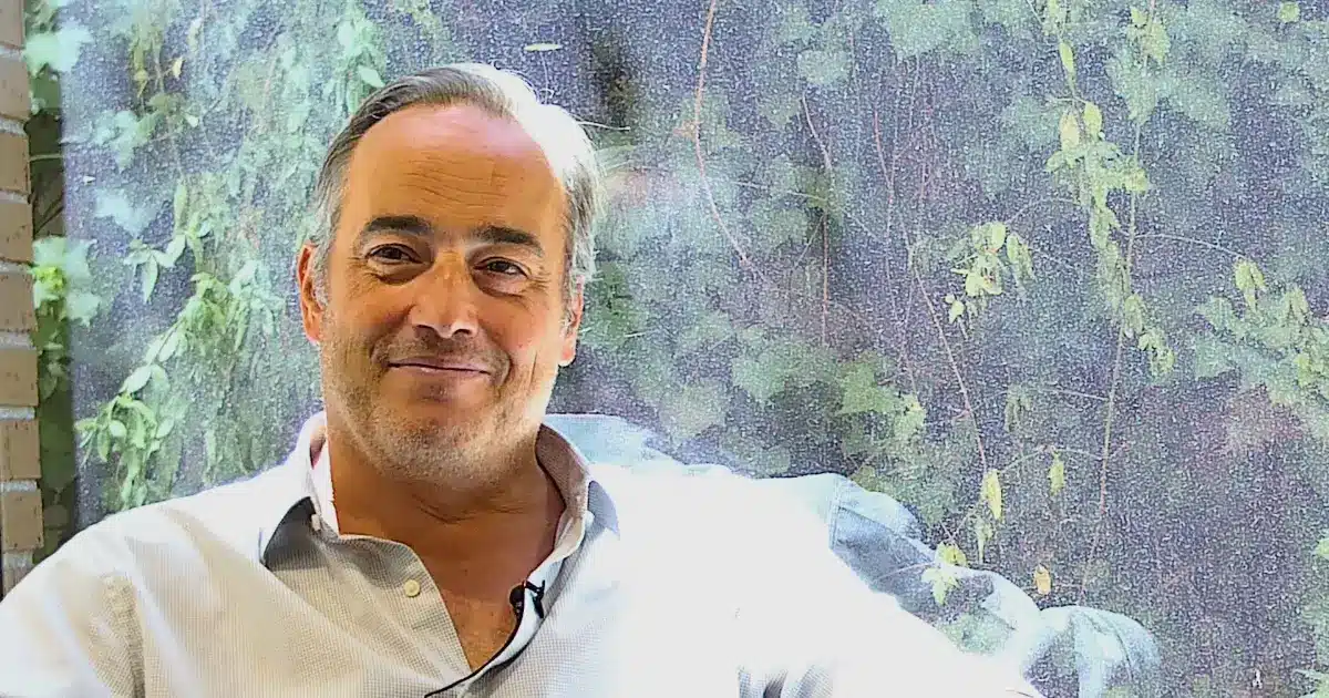 Retrato corporativo: hombre canoso sonriente con camisa celeste y micrófono de solapa, sentado junto a una ventana con enredaderas